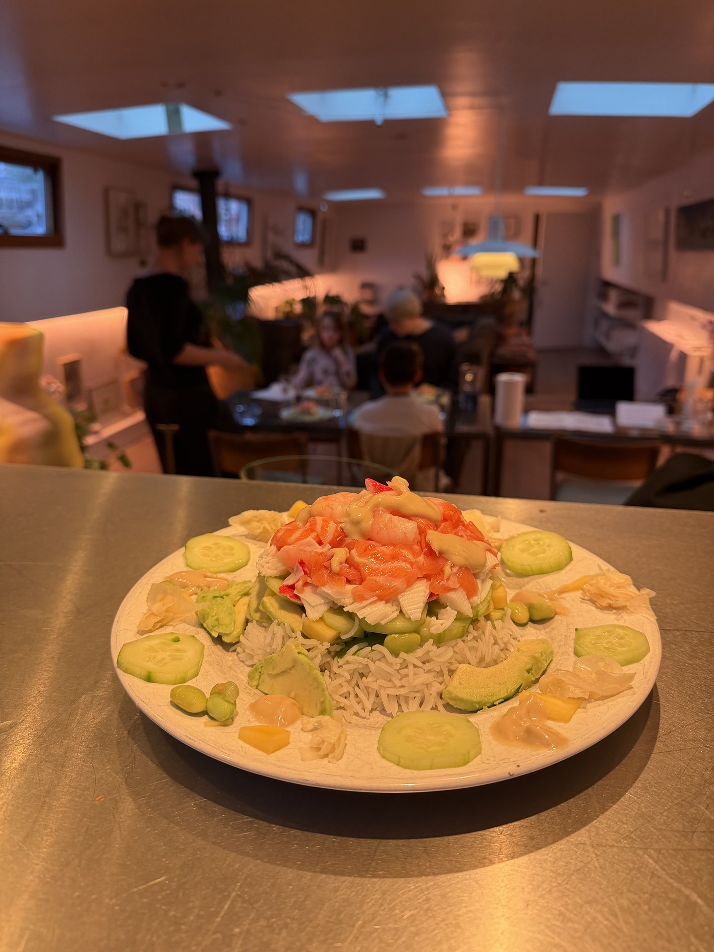 A vibrant seafood and vegetable rice bowl is prominently displayed in the foreground, featuring fresh ingredients like avocado, cucumber, and various seafood